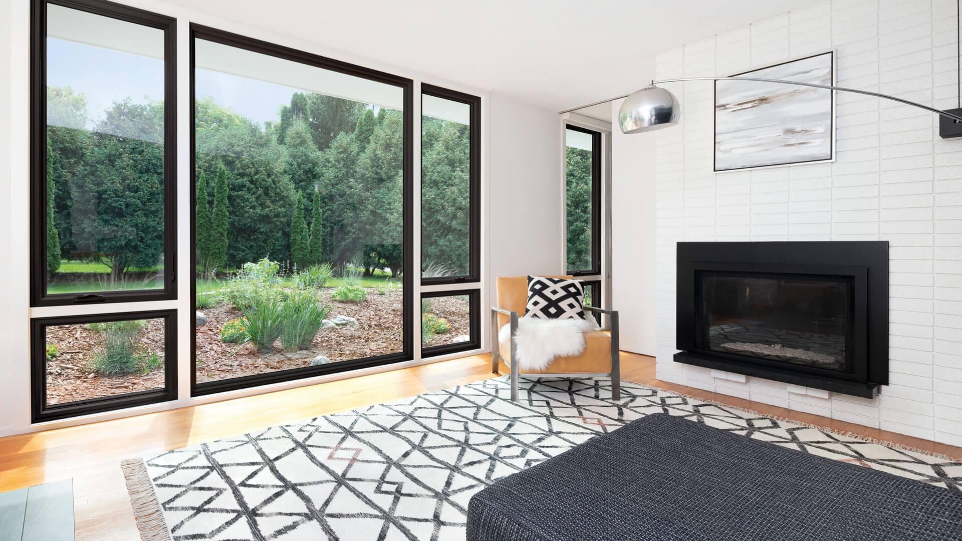 Spacious living room with wood flooring, white walls and modern fireplace, and five large picture windows overlooking green landscape.